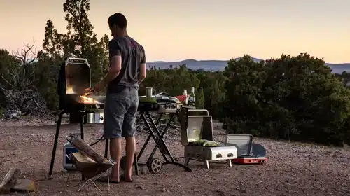 A backview of a person standing in fornt a grill at an evening setup at a campsite