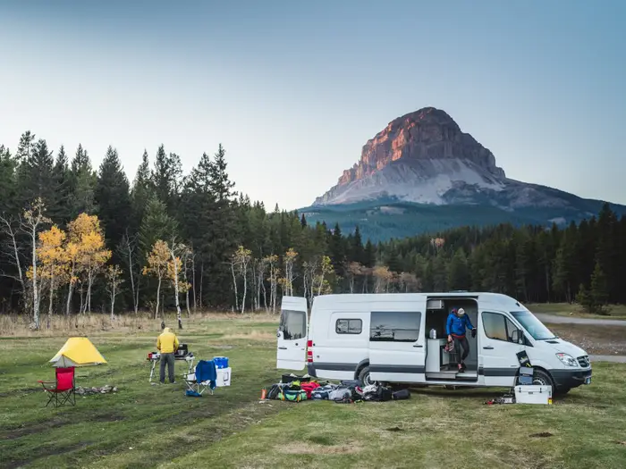 A beautiful view of a camping location with people setting up a camp alongisde a white camping van