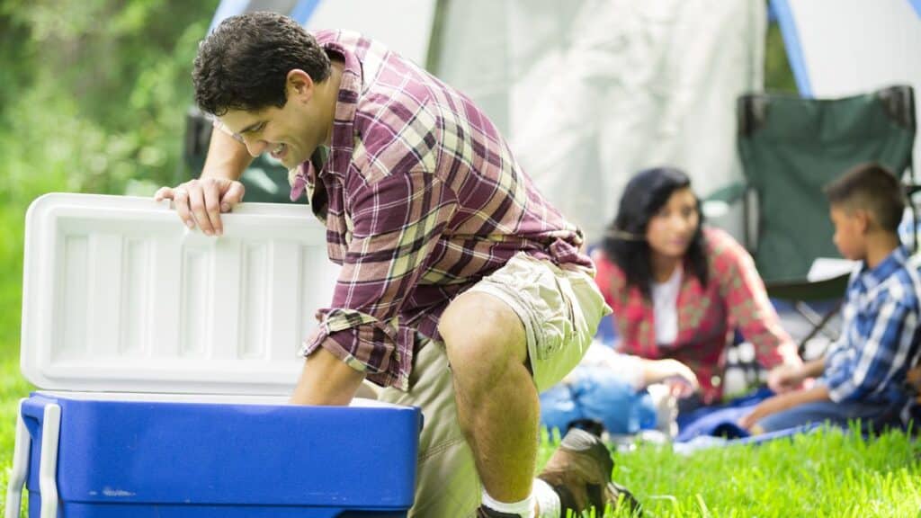 A man taking something out from a blue cooler with its family members sitting behind at a camping trip