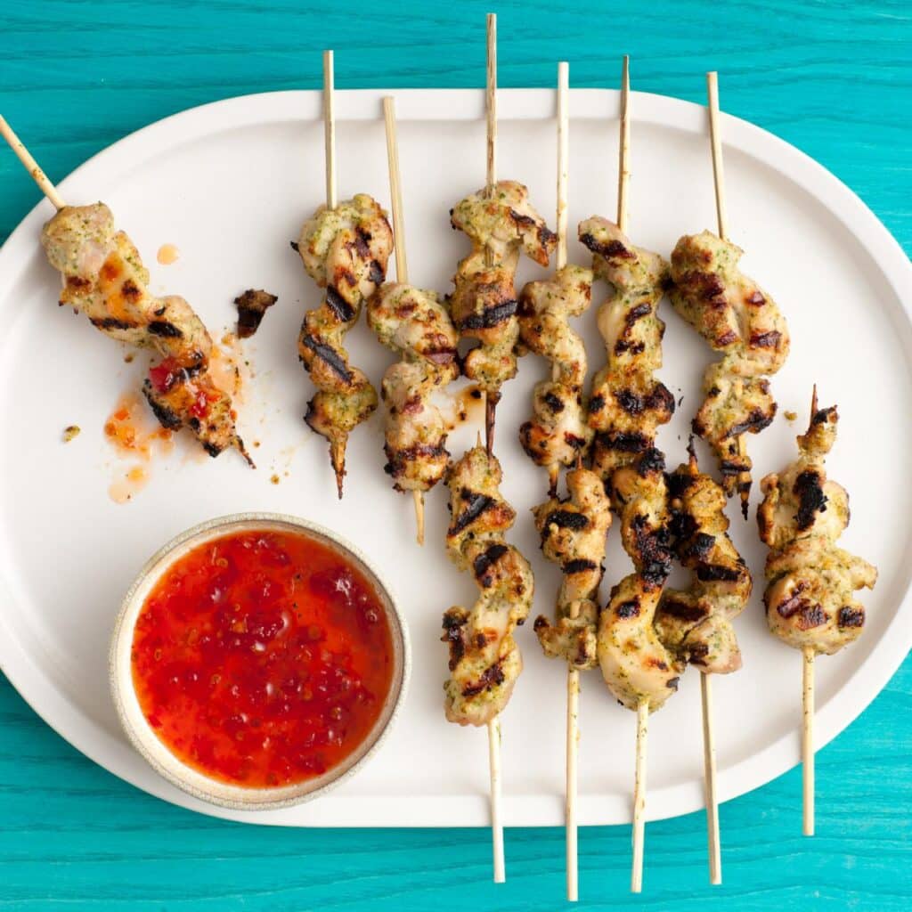 A top view of chicken on skewers plated out along with a bowl of chili sauce