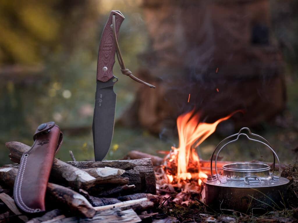 A view of a knife stuck inside a log beside a fire with a kettle at a campsite