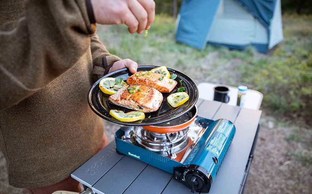 A view of a person garnishing fish with lemon beside an electric stove on a camp site