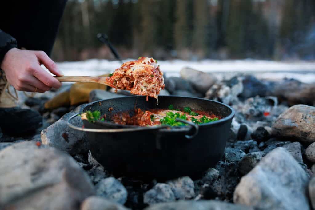 A view of a person taking out lasagna form a pan placed on the rocks at a campfire