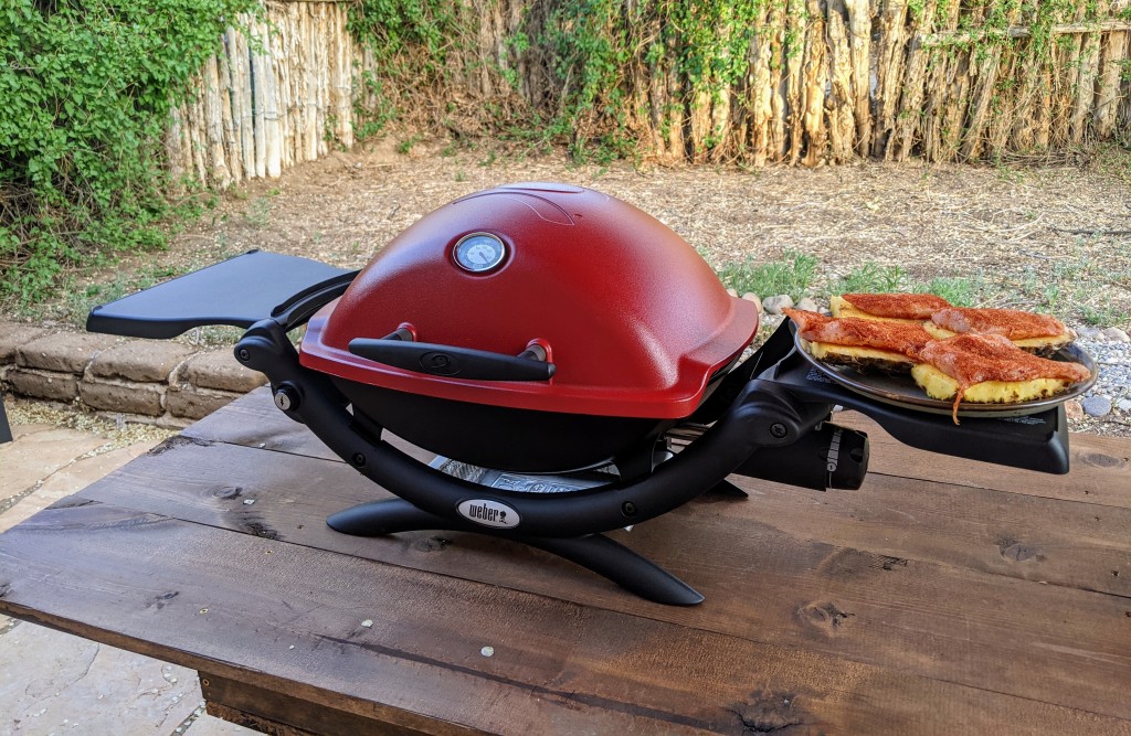 A view of a red Weber Q Camping Grill placed at a wooden bench alongside a meal