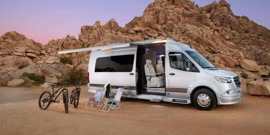 A view of a white camping van in a desert camp area with door opened and bicycles and chairs placed outside