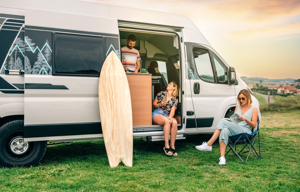 Two women sitting in front of camper van, one of them reading book while young man cooks