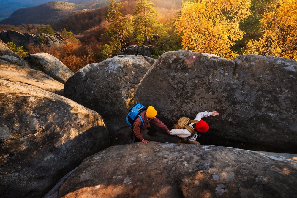 Two young tourists with backpacks are climbing between huge rocks to the top of mountain.