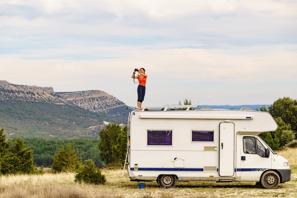 Woman enjoying trip with motor home, standing on roof of camper vehicle, holds camera, taking travel picture from mountain nature